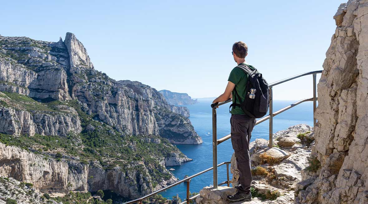 Man with backpack looking out across blue waters and white cliffs as he hikes a marked path with railings